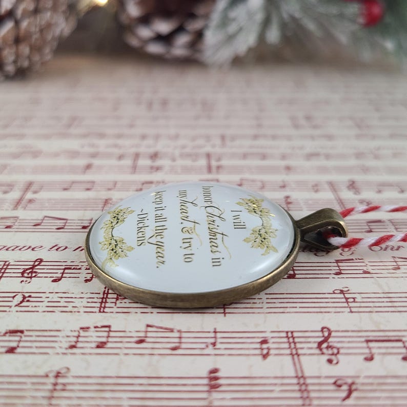 Pu&ograve; includere: Un ciondolo rotondo con una cornice in metallo color bronzo, sfondo bianco e testo dorato che recita: I will honor Christmas in my heart & try to keep it all the year. -Dickens. Il ciondolo &egrave; decorato con illustrazioni di agrifoglio dorato.