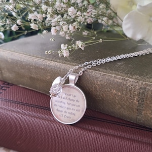 Puede incluir: Un collar con colgante plateado con un dije redondo que presenta una cita de C.S. Lewis. El dije se combina con un acento de cristal. El collar descansa sobre una pila de libros antiguos, con flores blancas en el fondo.