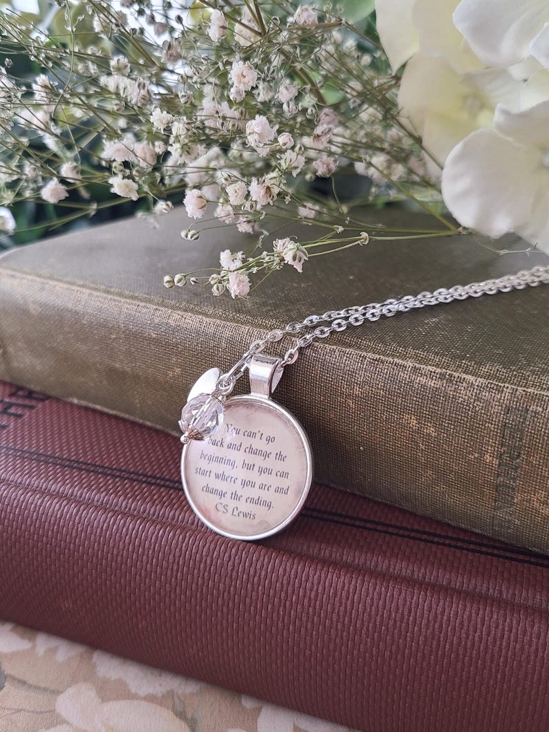 Puede incluir: Un collar con colgante plateado con un dije redondo que presenta una cita de C.S. Lewis. El dije se combina con un acento de cristal. El collar descansa sobre una pila de libros antiguos, con flores blancas en el fondo.