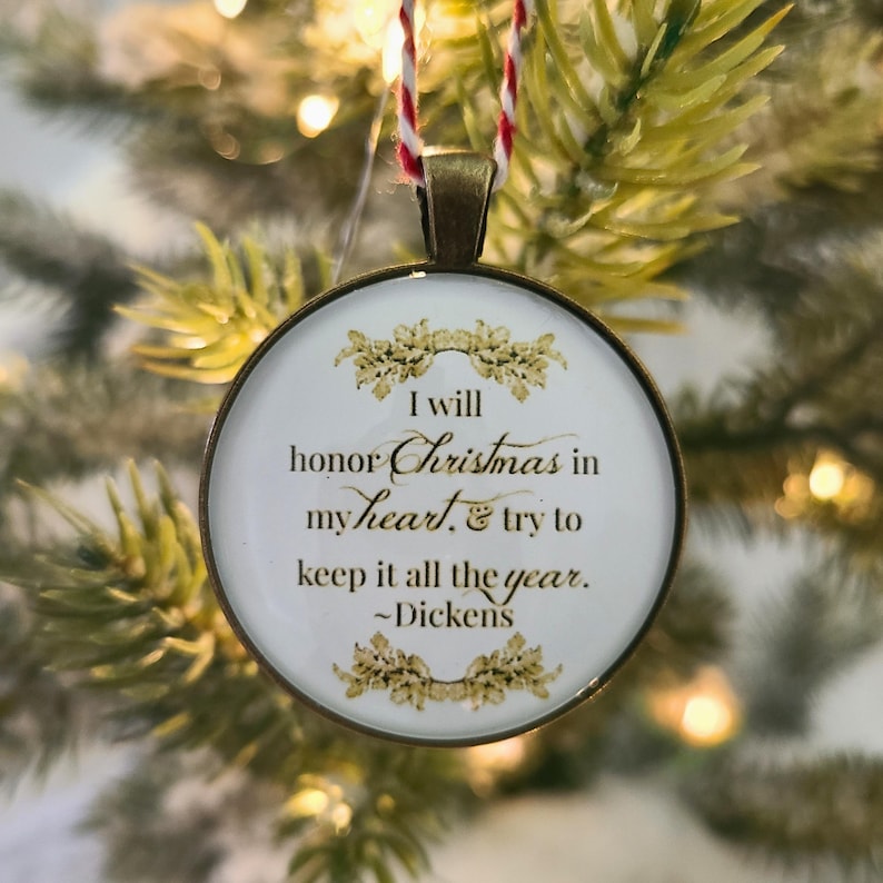 Pu&ograve; includere: Un ornamento rotondo color bronzo con sfondo bianco e testo dorato che recita: I will honor Christmas in my heart, & try to keep it all the year. ~Dickens. L'ornamento &egrave; appeso tramite un cordino ritorto di colore rosso e bianco.