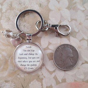Puede incluir: Llavero plateado con un colgante redondo con el nombre "Sarah" y una cita de C.S. Lewis. El llavero incluye un dije de coraz&oacute;n y una cuenta de cristal transparente. Una moneda estadounidense est&aacute; al lado.