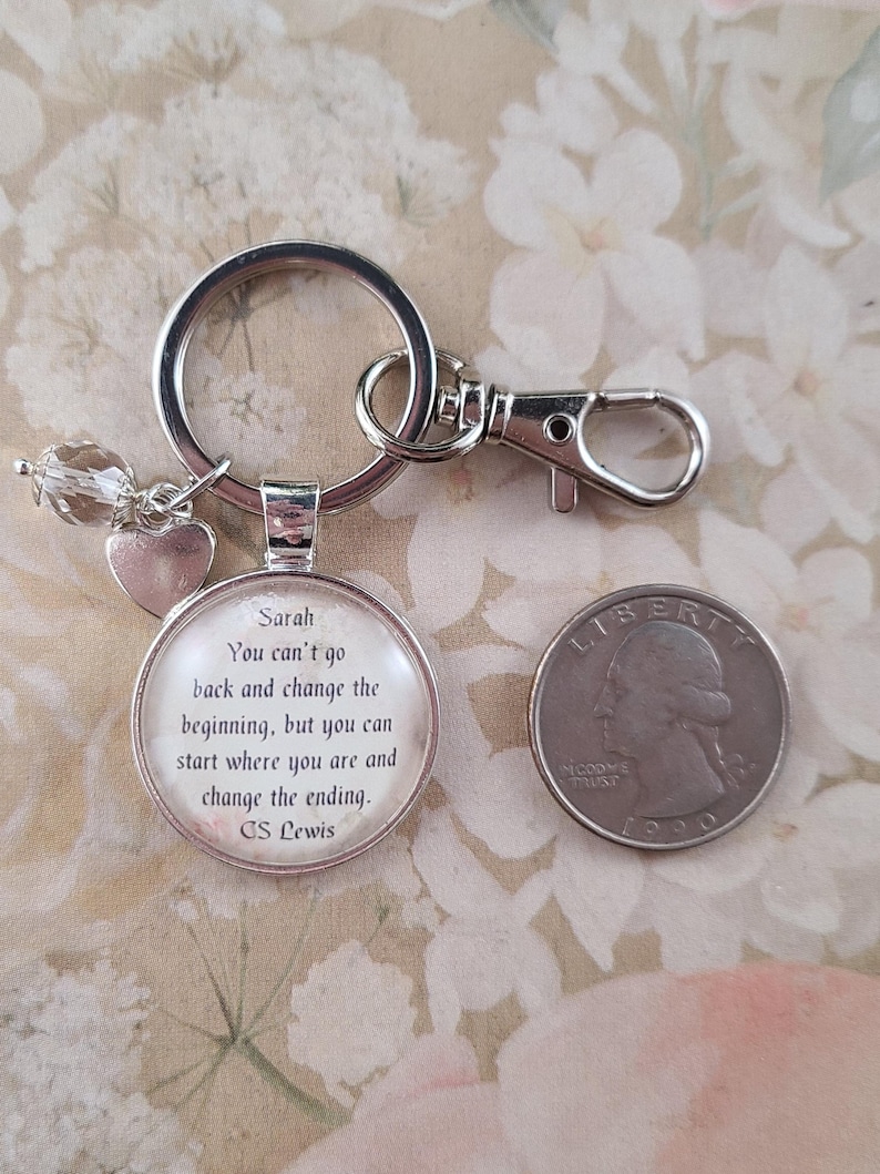 Puede incluir: Llavero plateado con un colgante redondo con el nombre "Sarah" y una cita de C.S. Lewis. El llavero incluye un dije de coraz&oacute;n y una cuenta de cristal transparente. Una moneda estadounidense est&aacute; al lado.