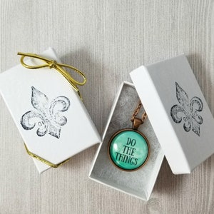 May include: A white gift box with a gold ribbon and a fleur-de-lis design. The box contains a necklace with a round glass pendant that says "Do the things" in black text on a teal background.