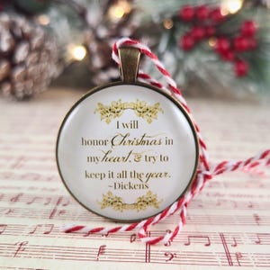 Pu&ograve; includere: Un ciondolo ornamentale rotondo color bronzo con sfondo bianco, decorazioni dorate e il testo: I will honor Christmas in my heart, & try to keep it all the year. ~Dickens. &Egrave; appeso a un cordino intrecciato di colore rosso e bianco.