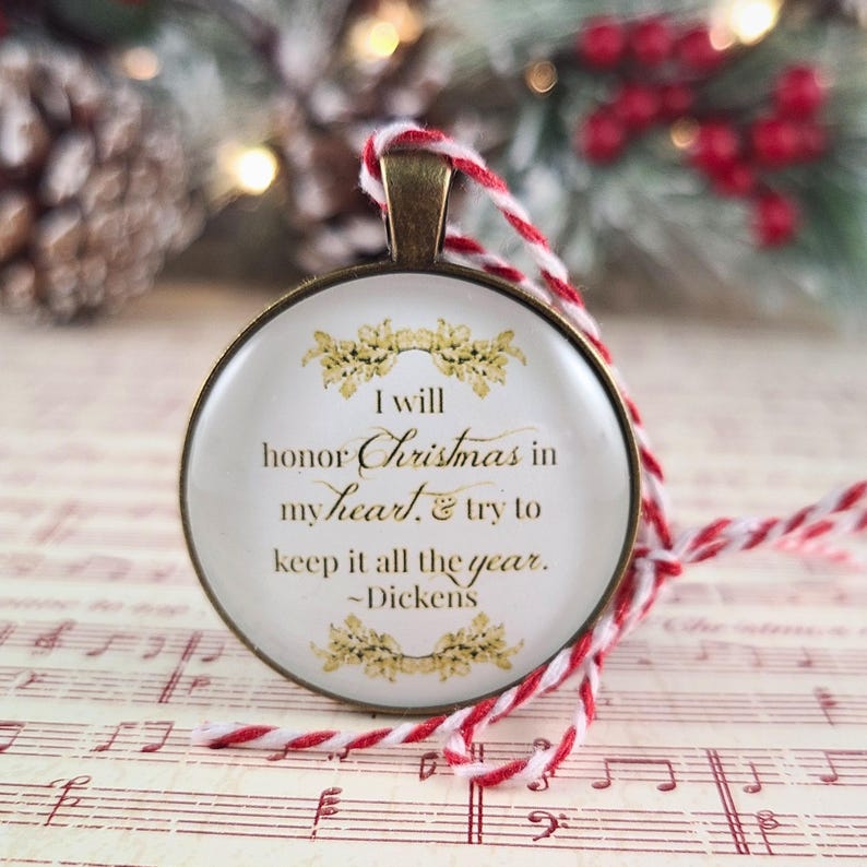 Pu&ograve; includere: Un ciondolo ornamentale rotondo color bronzo con sfondo bianco, decorazioni dorate e il testo: I will honor Christmas in my heart, & try to keep it all the year. ~Dickens. &Egrave; appeso a un cordino intrecciato di colore rosso e bianco.