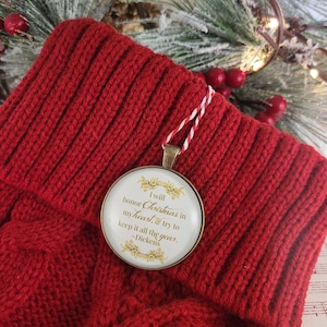 Pu&ograve; includere: Un ciondolo rotondo color bronzo con un inserto bianco e testo dorato che recita: I will honor Christmas in my heart & try to keep it all the year. -Dickens. Il ciondolo &egrave; appeso a un cordino intrecciato rosso e bianco su un tessuto a maglia rosso.