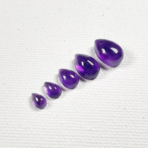 May include: A set of six amethyst gemstone cabochons in various sizes, all shaped like teardrops. The stones are a deep purple color and have a smooth, polished finish.