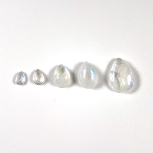 May include: Five teardrop-shaped moonstone cabochons of varying sizes, arranged in a row from smallest to largest on a white surface.