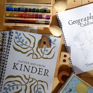 Może przedstawiać: Drewniany zestaw farb akwarelowych z różnymi kolorami, pędzlami i notatnikami. Na jednym notatniku widnieje napis "GATHER AND GROW KINDER", a na drugim "Geography Coloring". Obecne są również drewniane klocki z literami i drewniany przedmiot w kształcie jaja.