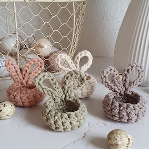 Könnte beinhalten: Drei gehäkelte Osternester in Beige, Grün und Braun in Form von Hasen. Die Nester stehen auf einer weißen Oberfläche mit Wachteleiern.