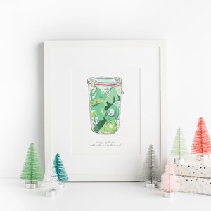 Op de afbeelding: Een wit ingelijste illustratie van een glazen pot gevuld met groene ingelegde augurken. De pot heeft een rood en wit deksel met een groen en wit etiket waarop staat "Ingelegde augurken met zout en mosterdzaad".