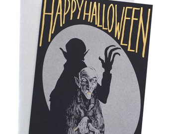 Puede incluir: Una tarjeta de Halloween negra y plateada con el texto "Happy Halloween" en letras doradas. La tarjeta presenta la silueta de un vampiro con colmillos y un vampiro más pequeño con colmillos frente a una luna llena.