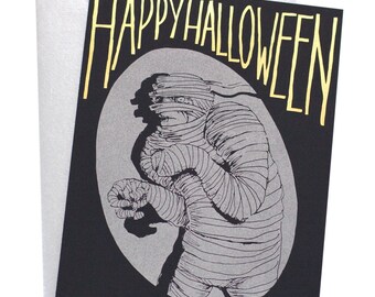 Puede incluir: Una tarjeta negra con letras doradas en relieve que dicen "Happy Halloween". La tarjeta presenta una ilustración en blanco y negro de una momie.