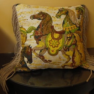 May include: A decorative pillow with a fringe border features a colorful design of four horses in a carousel setting. The horses are brown, yellow, green, and white with intricate details. The background is a cream color with a subtle pattern.