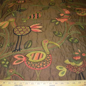 May include: Brown fabric with an embroidered design featuring colorful birds, leaves, and branches. The birds have orange, green, and brown details. The fabric has a textured appearance.