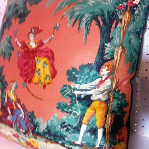 May include: A pink pillow with a colorful floral pattern featuring two figures in a whimsical scene. One figure is a woman in a red and yellow dress, and the other is a man in a blue and yellow outfit. The background includes green trees and a palm tree.