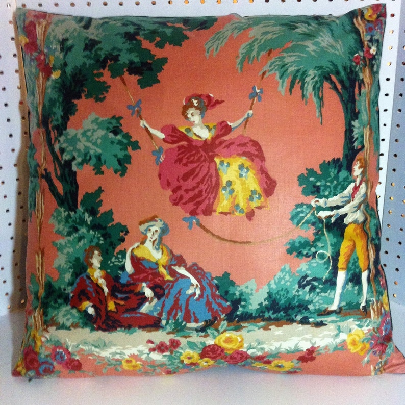 May include: A decorative pillow with a peach background and a colorful floral pattern. The pattern features a woman swinging on a rope, two people sitting on the ground, and a man standing on the right side of the pillow. The pillow has a green border.