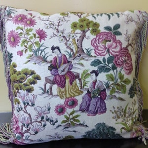 May include: A decorative pillow with a white background and a floral pattern featuring pink and purple flowers, green leaves, and two figures playing musical instruments. The pillow has a fringe border.