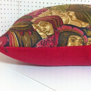 May include: A red and gold patterned pillow with a red velvet border. The pattern features a group of people in medieval clothing.