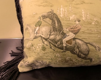 May include: A green pillow with a black fringe border features a vintage illustration of two riders on horseback. The riders are dressed in jockey attire and are riding through a field of green grass and trees.