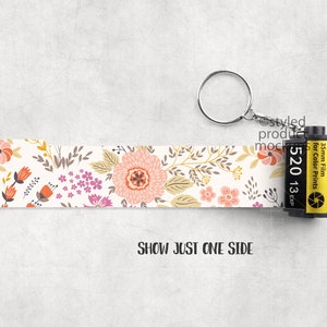 May include: A floral patterned keychain with a 35mm film canister. The keychain features a cream-colored strip adorned with pink, orange, and purple flowers and foliage. The film canister is black and yellow, with text that reads "520 13 EXP".