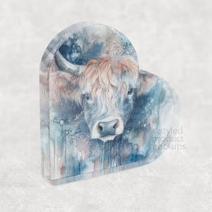 Pu&ograve; includere: Un oggetto decorativo a forma di cuore con un dipinto ad acquerello di una mucca delle Highlands. L'opera d'arte mostra il volto della mucca con pelo marrone e bianco, su uno sfondo di lavaggi ad acquerello blu e grigi. L'oggetto ha un aspetto strutturato.