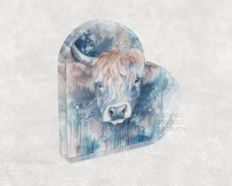 Pu&ograve; includere: Un oggetto decorativo a forma di cuore con un dipinto ad acquerello di una mucca delle Highlands. L'opera d'arte mostra il volto della mucca con pelo marrone e bianco, su uno sfondo di lavaggi ad acquerello blu e grigi. L'oggetto ha un aspetto strutturato.