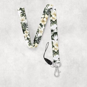 May include: A white lanyard with a floral pattern of cream-colored flowers and green leaves. The lanyard has a black clasp and a silver-colored metal clip for attaching keys or an ID badge.