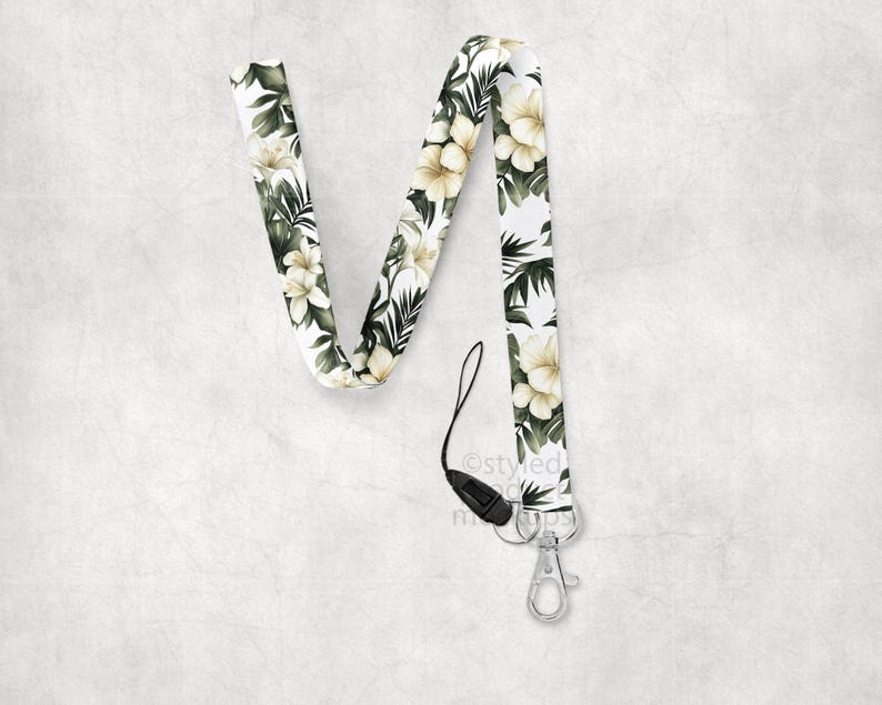 May include: A white lanyard with a floral pattern of cream-colored flowers and green leaves. The lanyard has a black clasp and a silver-colored metal clip for attaching keys or an ID badge.