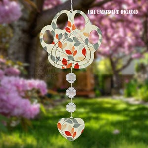 May include: A paw-shaped wind chime with a floral design in red, orange, and green. The wind chime hangs from a string and features clear, faceted crystals and a heart-shaped charm. The background is a blurred outdoor scene.
