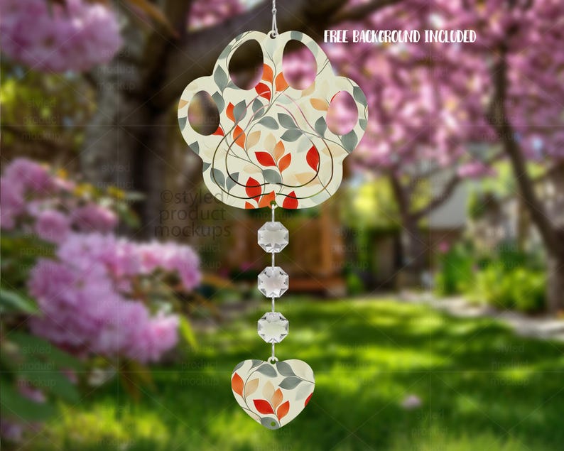 May include: A paw-shaped wind chime with a floral design in red, orange, and green. The wind chime hangs from a string and features clear, faceted crystals and a heart-shaped charm. The background is a blurred outdoor scene.