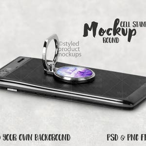 May include: A black smartphone with a silver phone ring holder attached. The ring holder has a round, colorful design with the words "Be the Magic" printed on it. The phone is lying on a gray surface. Cell Stand Mockup Round.