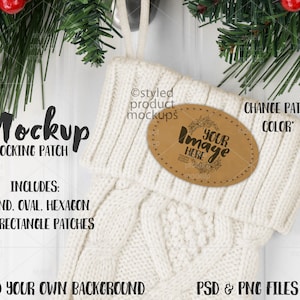 Op de afbeelding: Witte gebreide kous met een bruin leren patch waarop "Uw afbeelding hier" staat. De kous ligt op een witte houten achtergrond met groene dennentakken en rode bessen.