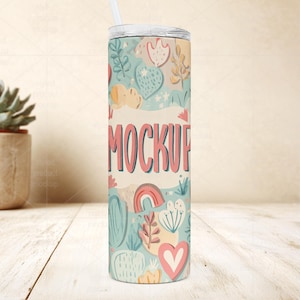 May include: A tall, insulated tumbler with a lid and straw. The tumbler is decorated with a whimsical pattern of hearts, rainbows, and botanical elements in pastel colors. The word "MOCKUP" is printed in pink across the center.