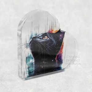 Pu&ograve; includere: Un blocco acrilico a forma di cuore con un design di gatto nero. Il gatto guarda in alto, con una rappresentazione dettagliata della pelliccia e dei baffi. Lo sfondo presenta una miscela di colori, tra cui blu, rosso e viola.