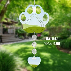 May include: A paw-shaped wind chime with a heart-shaped bottom, featuring a landscape design in shades of green and blue. The chime includes three crystal-like beads and the text "INCLUDES CANVA FRAME".