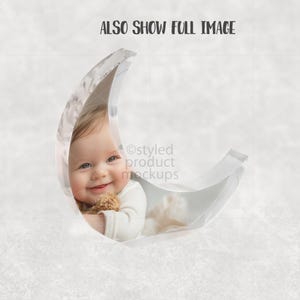 May include: A smiling baby in a white onesie is nestled inside a clear crescent moon prop. The baby is holding a small stuffed animal. The text "ALSO SHOW FULL IMAGE" is at the top of the image.