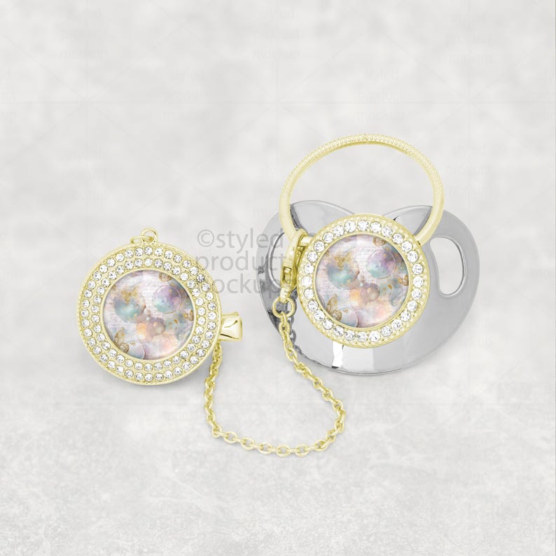 May include: A silver baby pacifier with a gold-colored ring and a matching clip. Both the pacifier and clip have a round, gold-colored center with a colorful design and a ring of clear crystals. A gold chain connects the clip to the pacifier.