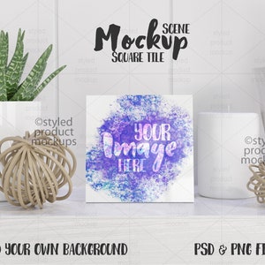 Könnte beinhalten: Ein weißer quadratischer Fliesen-Mockup mit einem Aquarell-Hintergrund. Die Fliese trägt den Text "YOUR IMAGE HERE" in lila und blauen Buchstaben. Die Fliese liegt auf einem weißen Regal mit einer weißen Kerze und einer dekorativen Kugel.