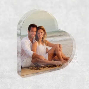 Pu&ograve; includere: Un blocco fotografico a forma di cuore in acrilico trasparente con una coppia seduta sulla spiaggia. L'immagine all'interno mostra un uomo e una donna in abiti bianchi, con uno sfondo di spiaggia e oceano.