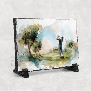 以下が含まれることがあります： 緑のゴルフコースでクラブを振るゴルファーの水彩画が描かれた装飾的なスレートプレート。アートワークには、木、青い空、草の生い茂る風景が含まれています。プレートは黒いスタンドで支えられています。