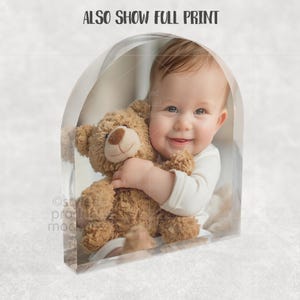 Op de afbeelding: Een lachende baby in een witte romper knuffelt een bruine teddybeer. De afbeelding is geplaatst in een helder, boogvormig frame, met de tekst "ALSO SHOW FULL PRINT" bovenaan. De achtergrond is zacht en neutraal.