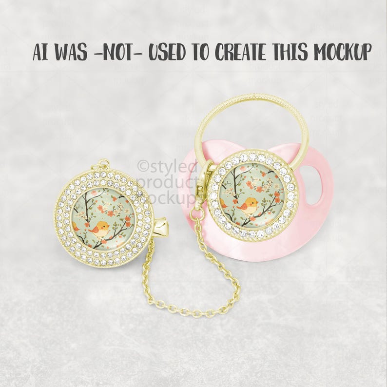 May include: A pink baby pacifier and matching pacifier clip. Both feature a gold-colored bezel encrusted with rhinestones. The center of each has a floral design with a small orange bird. The pacifier clip has a gold chain.