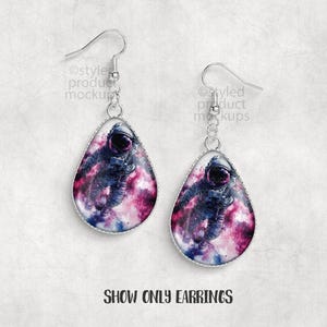 May include: A pair of silver-toned drop earrings. Each earring features a teardrop-shaped pendant with a space-themed design. The design shows an astronaut in a nebula of pink, purple, and blue hues. The text "SHOW ONLY EARRINGS" is at the bottom.