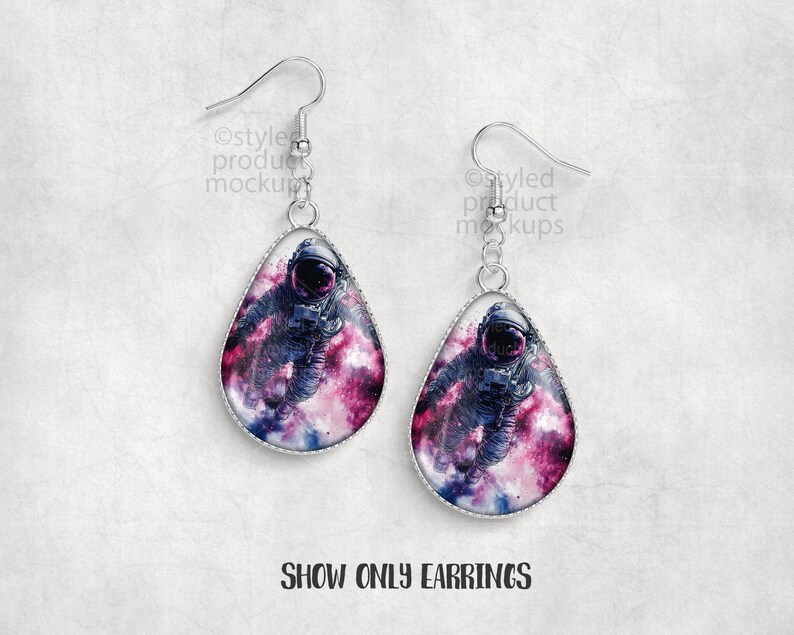 May include: A pair of silver-toned drop earrings. Each earring features a teardrop-shaped pendant with a space-themed design. The design shows an astronaut in a nebula of pink, purple, and blue hues. The text "SHOW ONLY EARRINGS" is at the bottom.