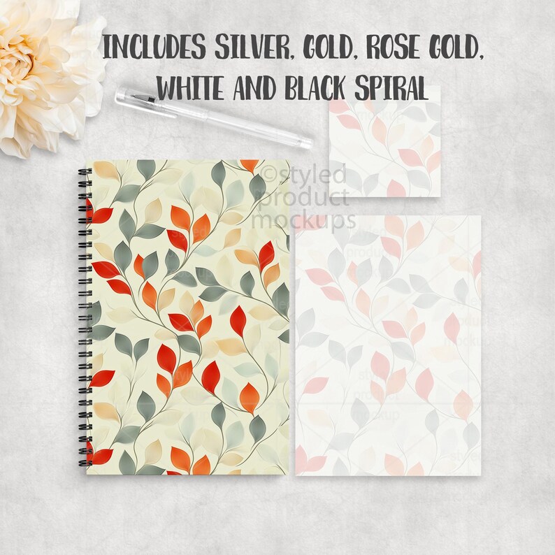 Op de afbeelding: Een spiraalgebonden notitieboek, een notitieblok en een kleine kaart met een bladpatroon in rood, oranje en grijs op een cr&egrave;mekleurige achtergrond. De tekst "INCLUDES SILVER, GOLD, ROSE GOLD, WHITE AND BLACK SPIRAL" staat bovenaan. Een pen is ook inbegrepen.