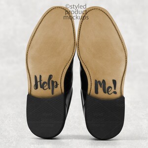 May include: A pair of black leather dress shoes with the soles facing up. The soles are tan with the words "Help Me!" written in black script. The shoes are on a light gray background.