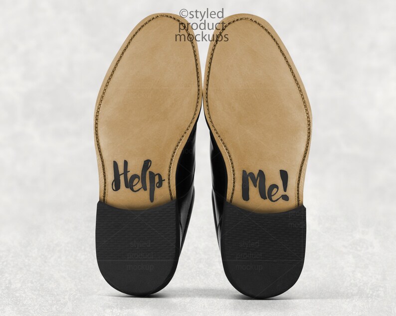 May include: A pair of black leather dress shoes with the soles facing up. The soles are tan with the words "Help Me!" written in black script. The shoes are on a light gray background.