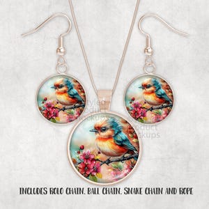 May include: A jewellery set comprising a pendant necklace and matching earrings. Each piece features a round, silver-toned frame with a colourful bird illustration perched on a flowering branch. The bird has blue, orange, and yellow feathers. The text in the image says "INCLUDES ROLO CHAIN, BALL CHAIN, SNAKE CHAIN AND ROPE."