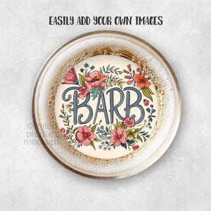 May include: A round coaster with the name "BARB" in blue script, surrounded by a floral wreath of red, pink, and yellow flowers with green leaves. The coaster is inside a clear glass with a bubbly liquid.